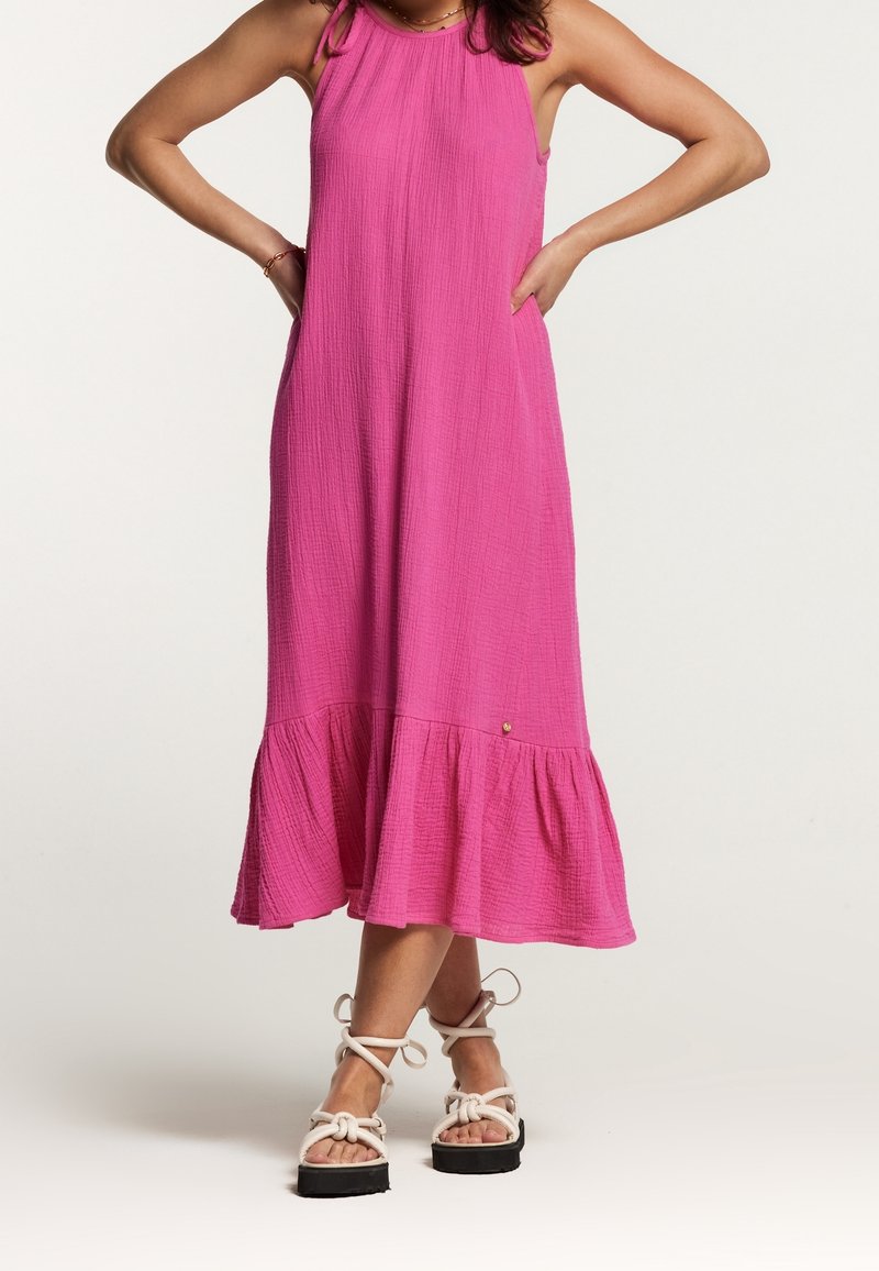 Femme en robe midi rose vif sans manches avec ourlet à volants, portant des sandales à plateforme blanches avec brides à la cheville, debout les mains sur les hanches.