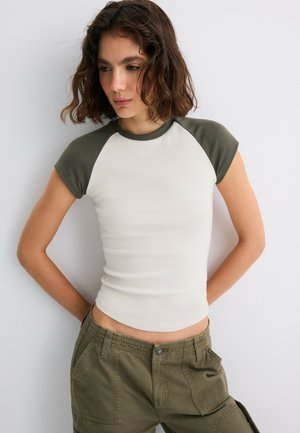 Ung kvinde med kort bølget hår ser ned, iført en hvid og olivengrøn kortærmet t-shirt og olivengrønne cargobukser med hænderne bag ryggen.