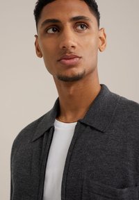Dunkelgraue Zip-Jacke mit Kragen, aus strukturiertem Stoff und mit einer Brusttasche, über einem weißen Hemd getragen. Minimalistisches Design.