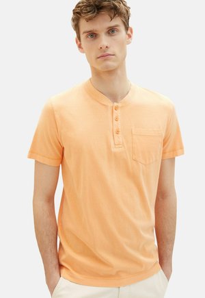 Camisa henley de manga corta, color naranja claro, con una tapeta de botones, bolsillo en el pecho y textura suave; diseño simple con un ajuste relajado.