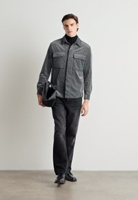 Gray striped button-up shirt with two chest pockets, paired with black pants and shoes. Model carries a black shoulder bag.