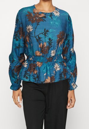 Frau trägt eine blaue, langärmelige Bluse mit Blumenmuster und geraffter Taille sowie schwarze Hose, steht vor weißem Hintergrund.