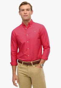 Czerwona koszula zapinana na guziki z długim rękawem, wykonana z bawełny, zestawiona z khaki spodniami. Charakteryzuje się plecionym brązowym paskiem i okrągłym zegarkiem na nadgarstku.