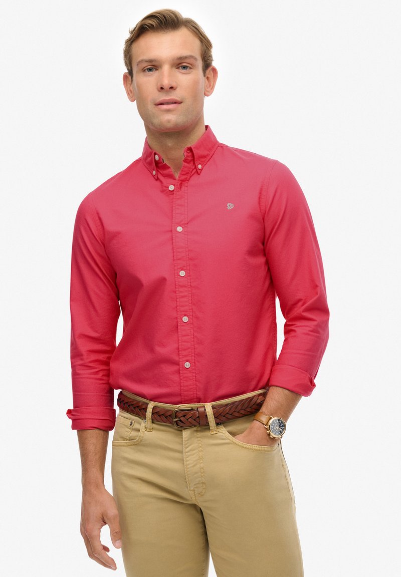 Czerwona koszula zapinana na guziki z długim rękawem, wykonana z bawełny, zestawiona z khaki spodniami. Charakteryzuje się plecionym brązowym paskiem i okrągłym zegarkiem na nadgarstku.