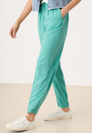 Person wearing teal drawstring pants with side pockets, white sneakers, and a blue denim vest, walking on a beige floor.