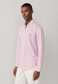 Homme portant une chemise à manches longues à rayures roses et blanches avec un logo brodé sur la poitrine, associée à un pantalon blanc, faisant face à l'avant.