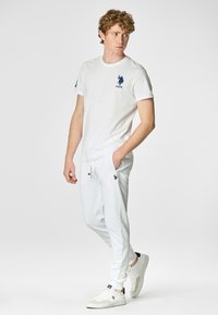 T-shirt bianca in cotone con logo blu marina sul petto, abbinata a pantaloni jogger bianchi con tasche laterali e logo ricamato, indossata con sneakers bianche.