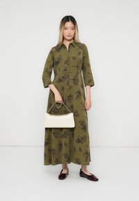 Donna che indossa un lungo vestito floreale verde e ballerine bordeaux, mentre tiene in mano una borsa bianca con tracolla a catena su uno sfondo semplice.