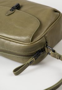Borsa in pelle verde oliva con chiusura con zip, forma rettangolare, tasca frontale piatta e tracolla staccabile. Superficie texture e accessori neri.