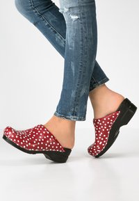 Zoccoli rossi con pois bianchi, realizzati in materiale lucido. Presentano una punta arrotondata, un retro aperto e una suola nera texturizzata. Indossati con jeans.