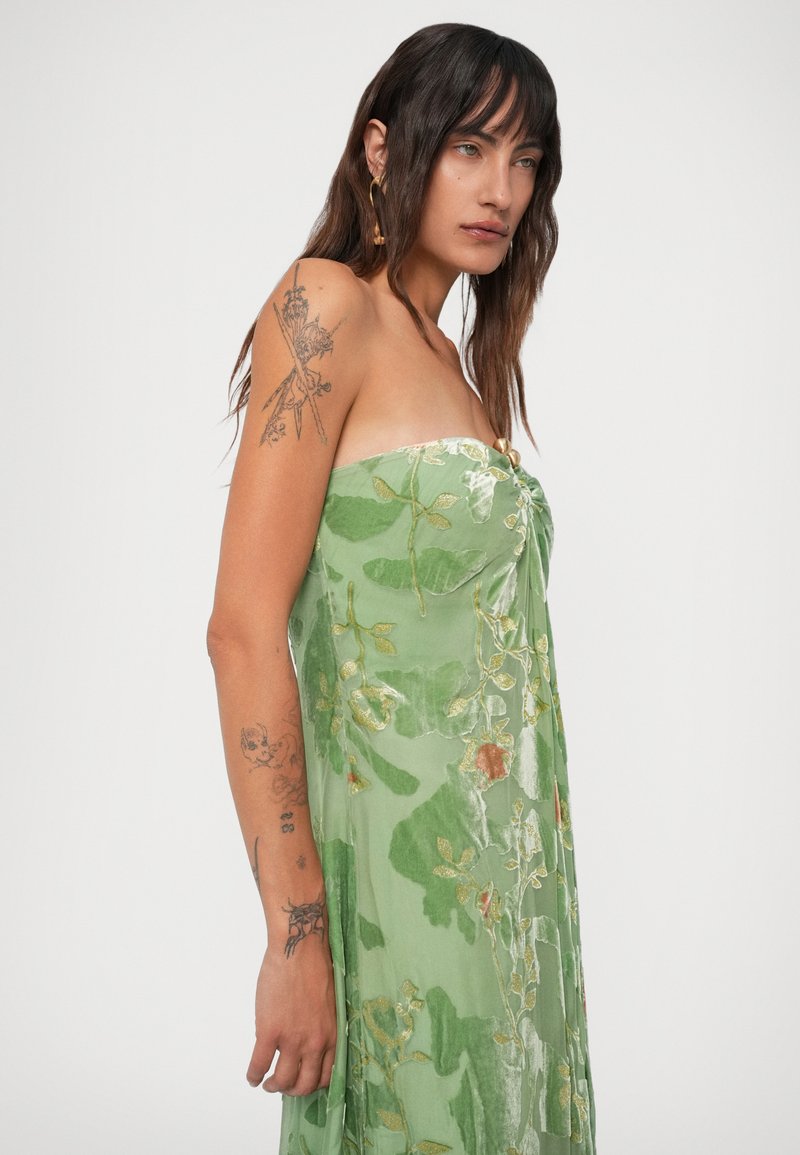 Woman with long dark hair, tattoos on left arm, wearing strapless green floral velvet dress with gold accents, standing against plain background.