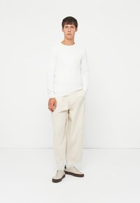Witte trui met lange mouwen en beige wijde broek, gecombineerd met lichte sneakers met donkerbruine accenten. Geplaatst tegen een effen achtergrond.