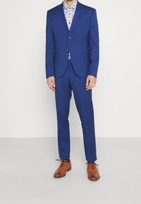 Costume bleu sur mesure avec un blazer ajusté, un pantalon assorti et une chemise à motif floral en dessous. Des chaussures habillées en cuir marron complètent le look.