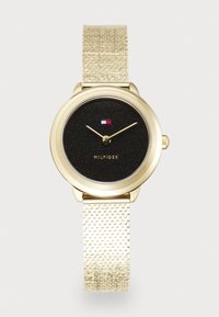 Montre dorée avec un bracelet en maille texturée, un cadran noir pailleté et des index horaires minimalistes. Affiche la marque "MILFGER" et un logo.