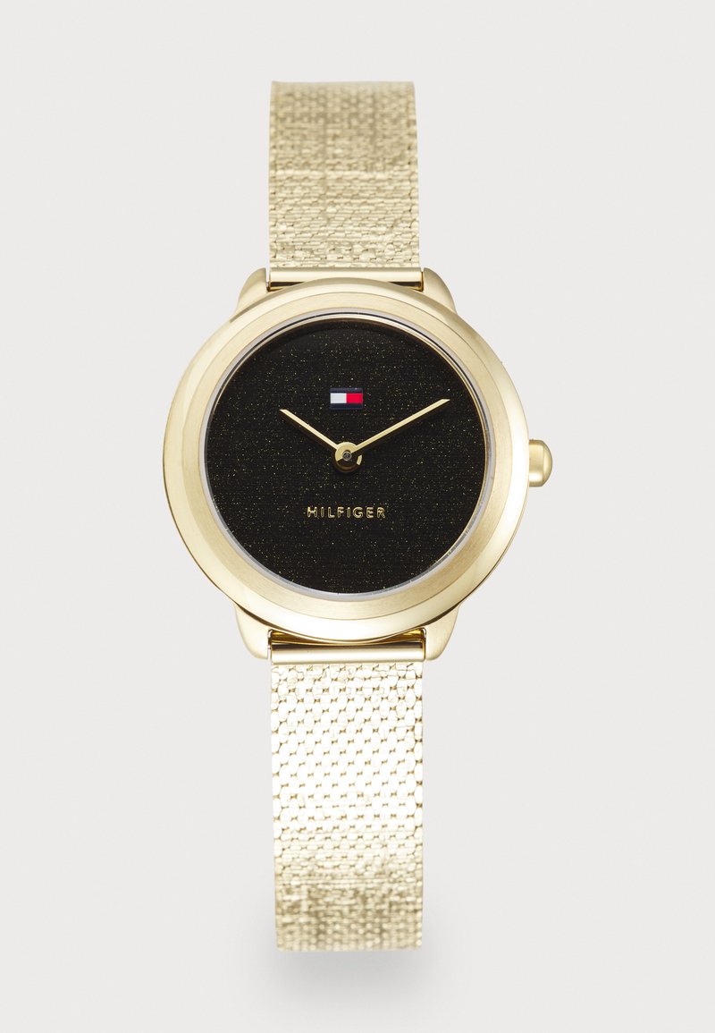 Zlatni sat s teksturiranom zlatnom mrežastom narukvicom, crnim sjajnim brojčanikom i minimalističkim oznakama sati. Sadrži "MILFGER" oznaku i logo.