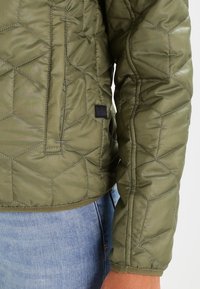 Olive green quilted jacket with hexagonal texture, featuring a black label and a fitted design, shown paired with blue denim jeans.