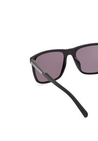 Mustat kulmikkaat aurinkolasit tummilla violetti linsseillä. Ominaisuuksia ovat sileä mattapinta ja metalliset yksityiskohdat temppeleissä.