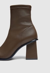 Bottine en faux cuir marron avec un haut talon carré. Présente un ajustement serré, des coutures discrètes et une texture lisse et brillante.