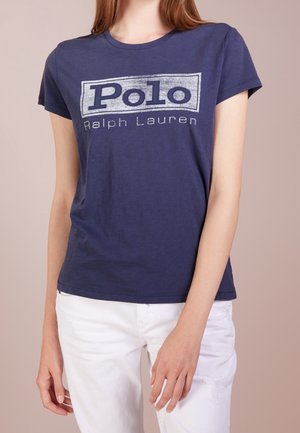 Kvinde iført en navy blå Polo Ralph Lauren t-shirt og hvide bukser, stående med sin venstre arm afslappet ned langs siden.