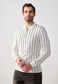 Polo shirt blanc à manches longues avec des rayures verticales navy, doté d'une patte zippée et de poignets côtelés, porté avec un jean foncé.