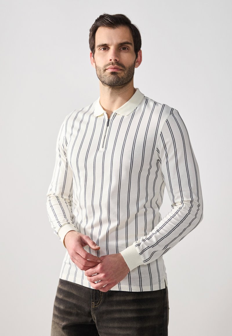 Polo shirt blanc à manches longues avec des rayures verticales navy, doté d'une patte zippée et de poignets côtelés, porté avec un jean foncé.