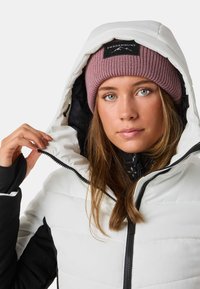Giacca piumino bianca con dettagli neri e chiusura con cerniera, dotata di cappuccio e foderata all'interno, abbinata a un cappello di lana rosa.