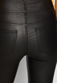 Černé kalhoty z umělé kůže s hladkým povrchem. Vysoký pas s dvěma zadními kapsami a viditelnými švy.