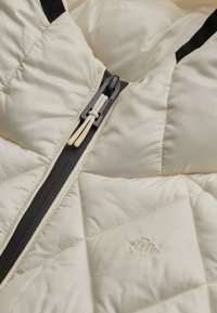 Beżowa pikowana kurtka puchowa z detalami: czarny zamek błyskawiczny, kremowe sznurki i wytłoczone logo po lewej stronie.