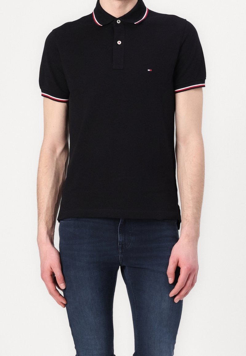 Homme portant un polo noir à manches courtes avec des bordures rouge et blanche sur le col et les manches, associé à un jean slim-fit bleu foncé.