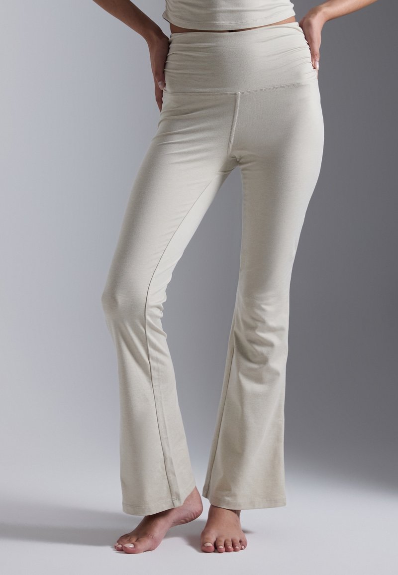 Femme portant un pantalon flare beige taille haute, pieds nus, debout devant un fond gris uni, les mains posées sur les hanches.