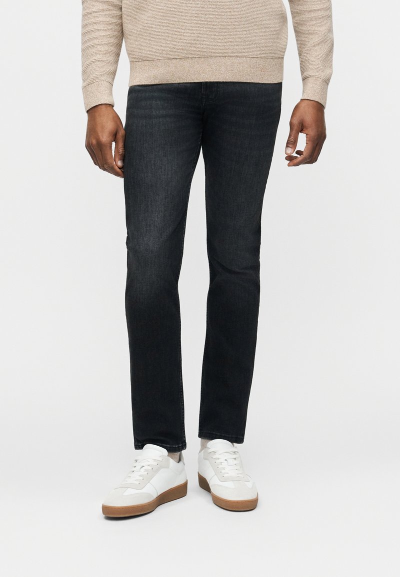 Άνδρας που φοράει μπεζ πουλόβερ, σκουρόχρωμα στενά τζιν και λευκά αθλητικά με καουτσούκ σόλες, στέκεται μπροστά σε απλό φόντο.