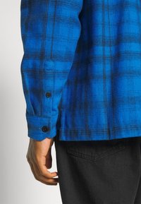 Blue plaid flannel shirt with black accents, featuring a buttoned cuff and textured fabric. Dark pants partially visible near the bottom.