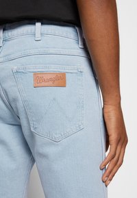 Los pantalones de mezclilla de color azul claro presentan un parche de cuero con el logo de Wrangler en el bolsillo trasero, con un diseño clásico de cinco bolsillos y costuras visibles.