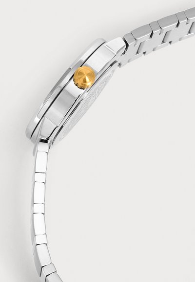 Vue latérale d'une montre-bracelet en métal argenté avec une couronne en or et un bracelet métallique lié sur fond blanc.