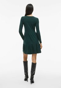 Robe tricotée à manches longues en vert foncé avec une texture côtelée, un corsage ajusté et une jupe évasée. Associée à des bottes noires à talons aiguilles jusqu'aux genoux.