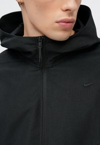 Czarna kurtka Nike z zamkiem z przodu, wysokim kołnierzem i teksturowaną tkaniną. Subtelne logo na piersi uzupełnia elegancki design.