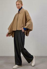 Veste cape beige avec col montant, design en couches et fermeture frontale, portée sur un col roulé bleu et un pantalon large à carreaux gris.