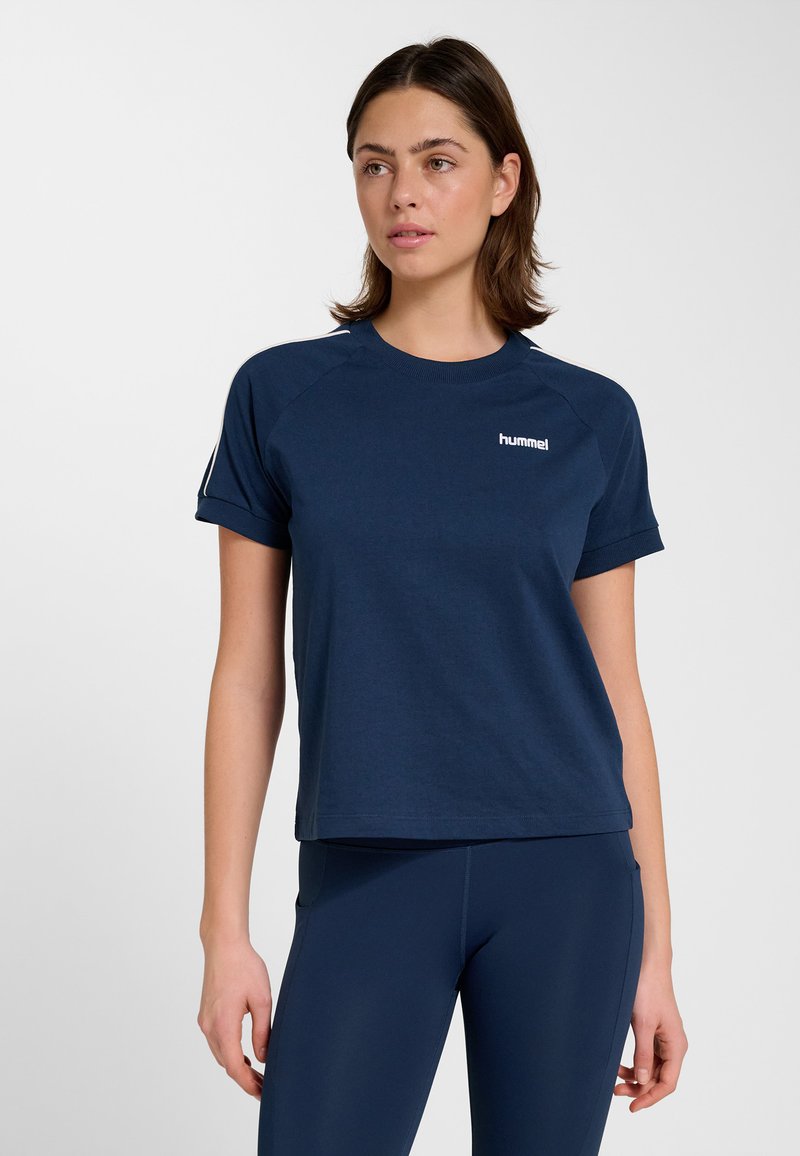 Femme portant un t-shirt hummel bleu marine à manches courtes et des leggings assortis, debout devant un fond clair uni.