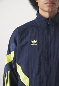 Marineblå vindjakke med gule detaljer, frontlynlås og Adidas-logo. Lavet af letvægts, struktureret stof med farveblok design.