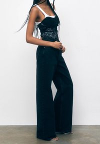 Top de corsé de encaje negro sobre una camiseta blanca, combinado con jeans oscuros de pierna ancha de cintura alta. La modelo lleva sandalias de punta abierta.