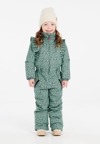 Groen patroon sneeuwpak met zwarte vlekken, met een hoge hals en ruffles, gecombineerd met een crèmekleurige gebreide muts en beige laarzen.