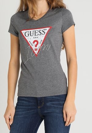 T-shirt en coton gris à manches courtes, avec un graphique triangulaire en rouge, noir et blanc, portant les inscriptions "GUESS" et "I'm" en texte décoratif.