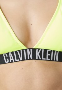Neon žlutý trojúhelníkový bralet s černým elastickým páskem, na kterém je bíle napsáno "CALVIN KLEIN". Hladká textura, minimalistický design.