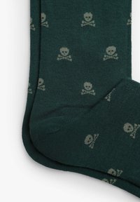Mørkegrønne sokker lavet af blødt stof, med et gentagende mønster af kranier og korslagte knogler i en lysere beige farve.