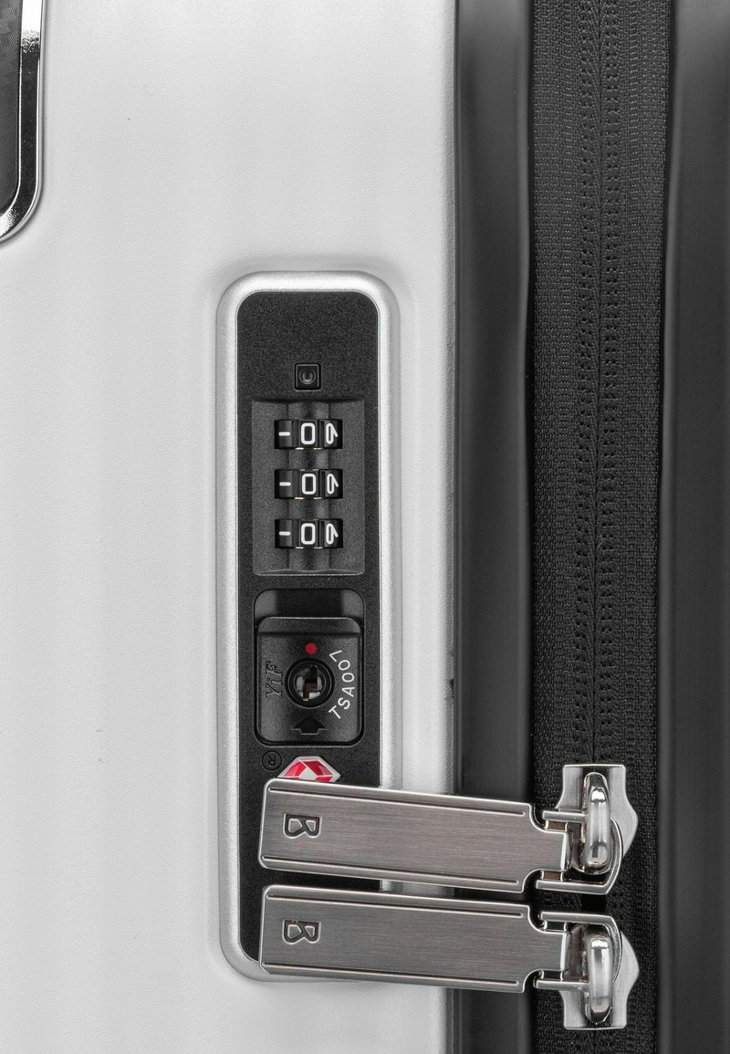 Biała walizka z rogu, wyposażona w zamek szyfrowy z numerycznymi tarczami, komorą na klucz TSA oraz srebrnymi zamkami na czarnym taśmie zamka.