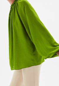 Blusa verde con colletto alto, dettaglio a volant arricciato e maniche lunghe a sbuffo. Realizzata in tessuto leggero, con una texture liscia.