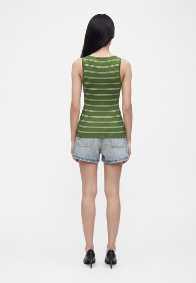 Femme aux longs cheveux noirs portant un débardeur rayé vert, un short en denim clair et des chaussures noires à talons hauts, debout, dos tourné.