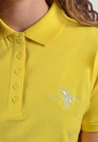 Gul pikétröja i texturerad bomull, med klassisk krage, sex knappar framtill och en diskret broderad logotyp på bröstet.