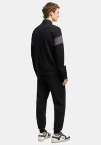 Tuta nera con una striscia viola sulle spalle, composta da giacca con zip e polsini elasticizzati, abbinata a sneakers bianche e nere.