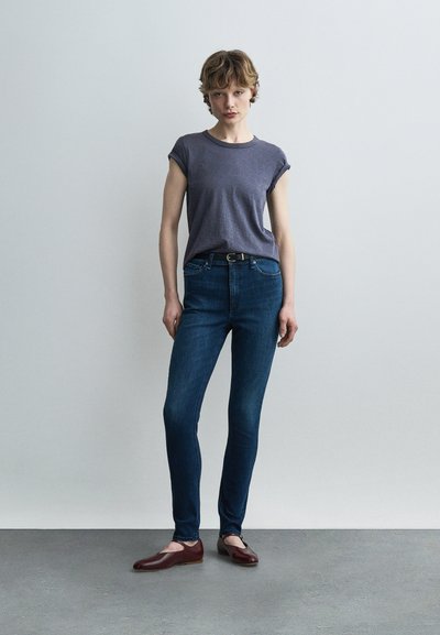 Νεαρό άτομο με κοντά μαλλιά που στέκεται, φοράει σκούρο γκρι T-shirt με γυρισμένα μανίκια, σκούρο μπλε στενά τζιν, μαύρη ζώνη και καφέ μπαλαρίνες.